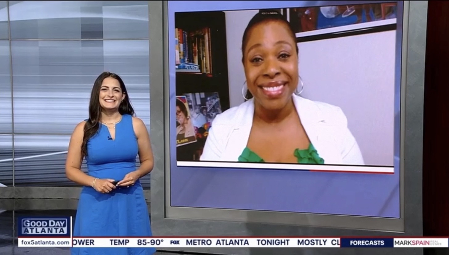 A news broadcast showing a smiling woman in a white jacket and a female news anchor in a blue dress standing in a studio.
