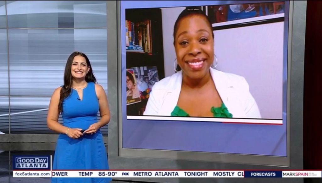 A news broadcast showing a smiling woman in a white jacket and a female news anchor in a blue dress standing in a studio.