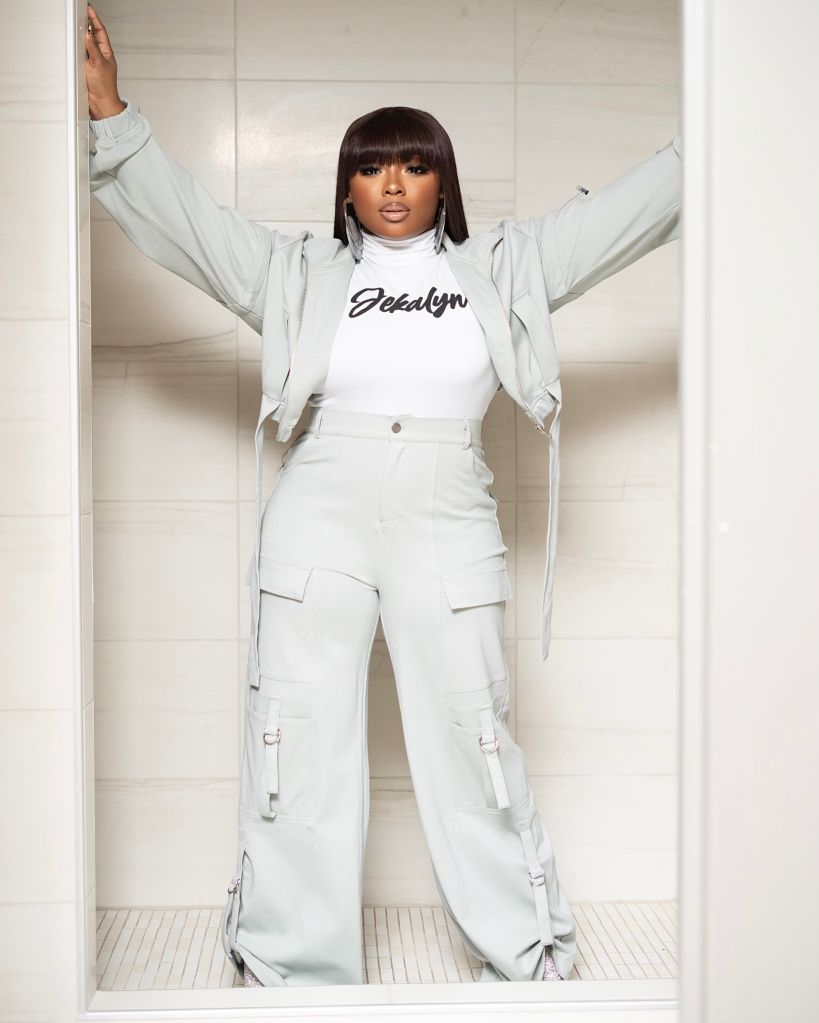 A woman in a white outfit with the text "Jekalyn" on her shirt, standing with her arms raised in a white room.