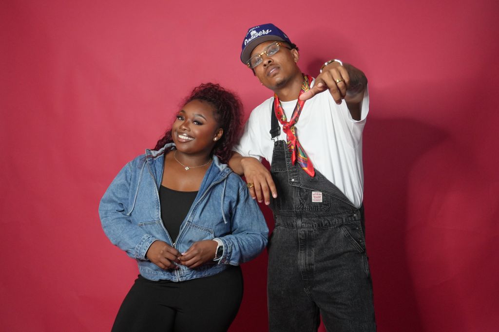Two people, a woman and a man, posing together against a red background. The man is wearing a baseball cap and the woman is wearing a denim jacket.