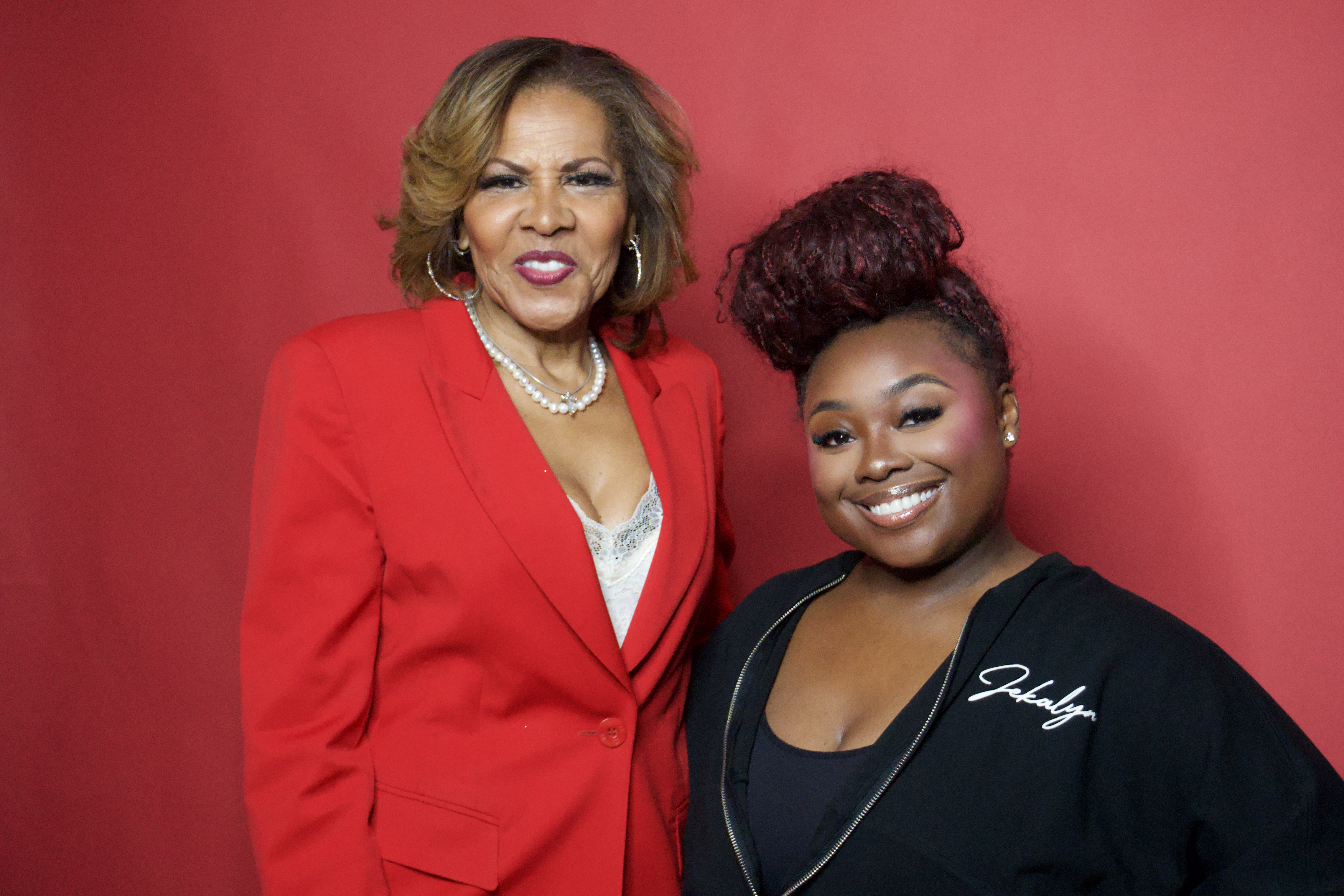 Two women posing together, one wearing a red jacket and the other a black top, both smiling against a red background.