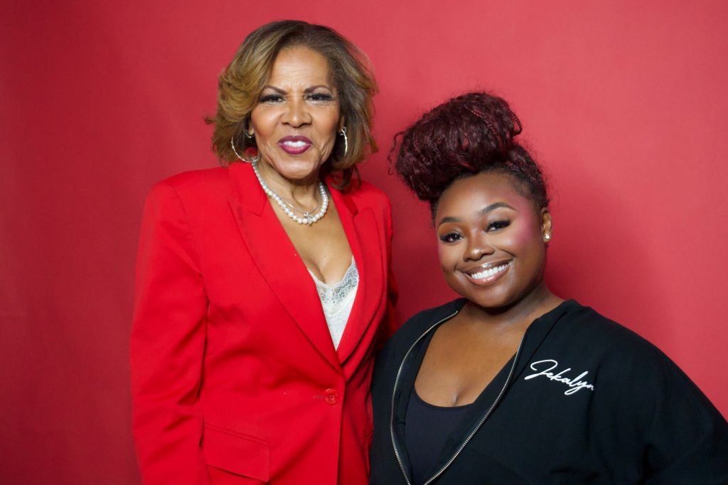 Two women posing together, one wearing a red jacket and the other a black top, both smiling against a red background.