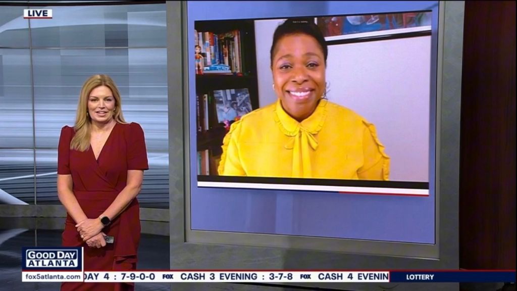 A news anchor in a red dress stands in a studio, with a smiling woman in a yellow coat shown on a screen behind her.